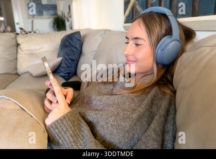 Schöne junge Frau mit Kopfhörern, die ihr Smartphone auf dem Sofa zu Hause benutzt. Technologiekonzept. Stockfoto