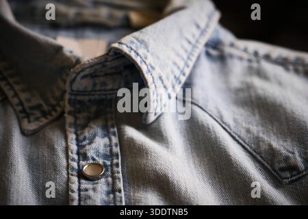 Nahaufnahme des hellblauen Denim-Hemdes mit Schwerpunkt auf Kragen, Nähten und Druckknopf aus Metall. Materialstruktur, Nähte und lässige Bekleidungsdetails Stockfoto
