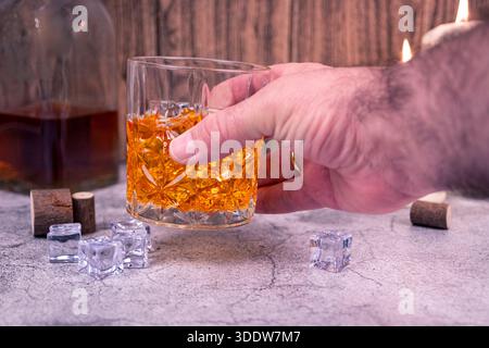 Hand hält ein geschliffenes Kristallglas mit Whiskey und Eiswürfeln auf einer strukturierten Oberfläche, Flasche im Hintergrund. Klassische Spirituosen und Barkonzept. Stockfoto