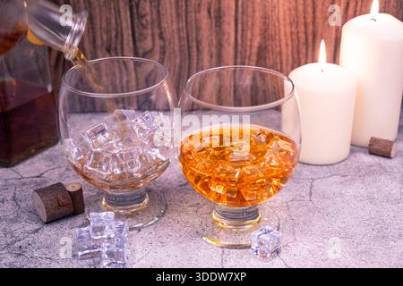 Whisky wird in einen mit Eis gefüllten Schnüffler gegossen, neben einem gefüllten Glas. Rustikaler Hintergrund mit Kerzenlicht, Bar und Suchtkonzept. Stockfoto