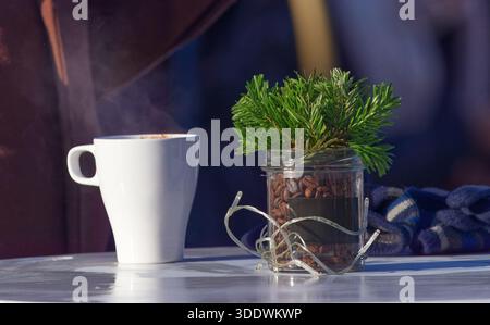 Warme, einladende Outdoor-Szene mit dampfend weißem Kaffeetasse neben einem Glas mit Bohnen und immergrünen Zweigen. Ideal für gemütliche Morgenstunden, W Stockfoto