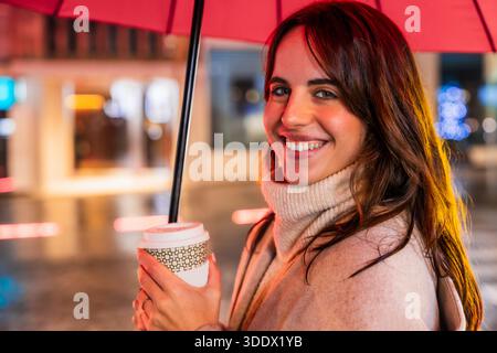 Lächelnde Frau, die nachts Kaffee unter rotem Schirm hält Stockfoto
