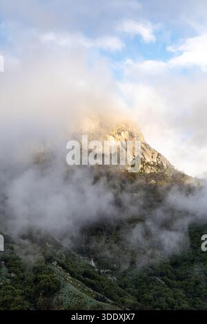 Dramatische Berglandschaft mit felsigem Gipfel, der über Morgennebel und niedrigen Wolken aufsteigt. Stimmungsvolle Naturszene mit sanftem Licht, nebeligem Wald und Friedhof Stockfoto
