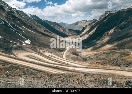 Gewundene Feldstraße durch einen Bergpass mit Felsen und schneebedeckten Gipfeln unter einem bewölkten Himmel. Reisen Sie durch raues Gelände für Abenteuerreisen Stockfoto