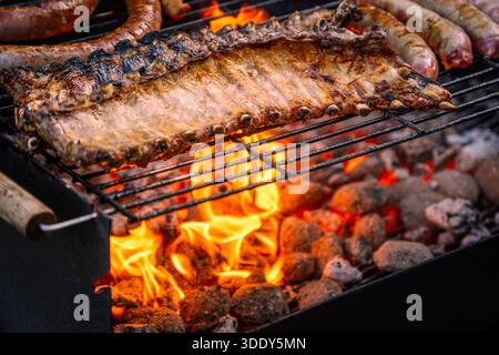 Nahaufnahme marinierter Schweinerippchen und Würstchen, die über intensiven Flammen und glühenden heißen Kohlen auf einem Grill grillen. Stockfoto