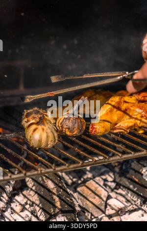 Eine Nahaufnahme einer Hand, die eine Zange hält, um gegrillte Hähnchenschenkel und eine ganze Knoblauchzehe über Holzkohlen zu bewegen. Stockfoto
