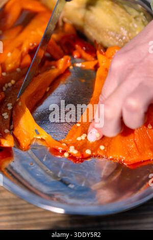 Vertikale Nahaufnahme von Streifen gerösteter Orangenpaprika, geschnitten auf einem runden reflektierenden Metallblech von den Händen einer Frau mit einem Messer. Stockfoto
