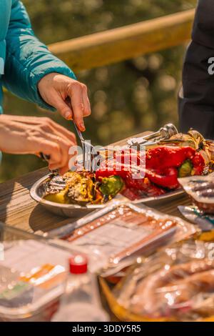 Eine Frau schneidet geröstetes Gemüse, Auberginen und rote Paprika mit Messer und Gabel auf einem Holztisch voll mit Fleisch und Saucen, um sie auf einem zu rösten Stockfoto