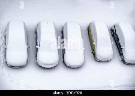 Blick von oben auf eine Reihe geparkter Autos, die komplett von Neuschnee bedeckt sind. Minimalistische Winterkomposition mit starker Geometrie, Wiederholung und sauberer weißer Kopie Stockfoto