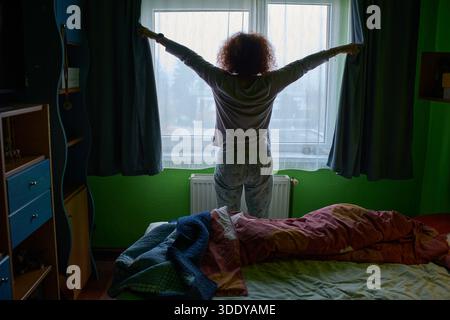 Frau im Schlafanzug öffnet die Vorhänge des Schlafzimmers, um nach dem Aufwachen das Morgenlicht einzulassen Stockfoto