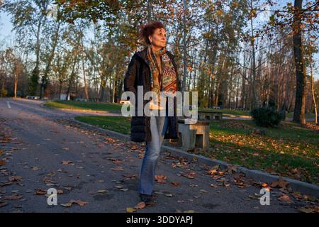 Stilvolle Frau in pufferfarbenem Mantel und farbenfrohem Schal, die auf dem Herbstparkweg mit gefallenen Blättern spaziert Stockfoto