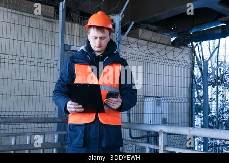 Männlicher Arbeiter in Sicherheitsausrüstung hält das Klemmbrett in der einen Hand und überprüft das Smartphone in der anderen in einem umzäunten Industriebereich im Freien Stockfoto