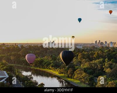 Fünf einzigartige Heißluftballons schweben über einem üppigen Flusspark und der Skyline der Stadt während eines ruhigen, goldenen Aufstiegs. Stockfoto