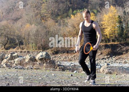 Sportlicher Mann wandert selbstbewusst auf einem Kiesweg im Freien, der einen orangen Badmintonschläger hält, mit Fluss und bewaldetem Hintergrund im Herbst. Stockfoto