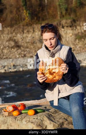 Nahaufnahme einer Frau, die einen Happen von warmen Fladenbrot bei einem malerischen Picknick am Fluss genießt, umgeben von farbenfrohem frischem Gemüse auf einer Holzoberfläche. Stockfoto