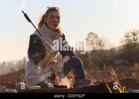Fokussierter Schuss einer Frau mit Pferdeschwanz, die einen weißen Marshmallow über orangefarbenen Lagerfeuer-Flammen röstet, in Weste gekleidet unter verschwommenem Herbstlaub (102) Stockfoto
