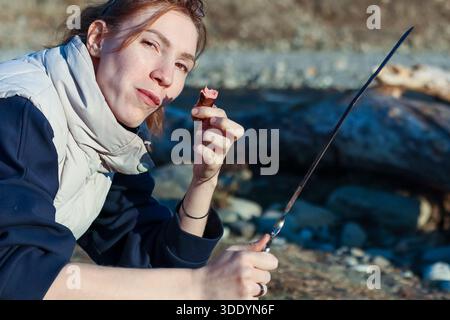 Nahaufnahme einer lässigen Frau in Winterkleidung, die geröstetes Fleisch auf einem Metallspieß inmitten von Flussfelsen röstet und schmeckt. Stockfoto