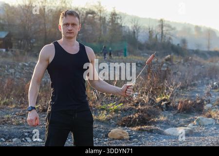 Empowered Outdoor Porträt: Muskulöser Mann in Fitnesskleidung posiert mutig mit Spießen gegrillter Wurst, hoch stehend in rauer Herbstlandschaft mit d Stockfoto