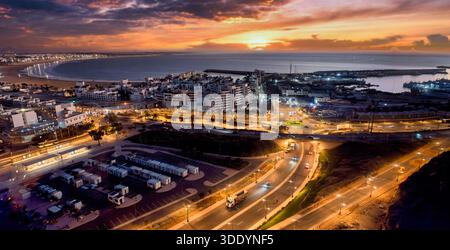 Eine lebhafte Küstenstadt bei Sonnenuntergang mit einem sichelförmigen Strand, beleuchtetem Hafen, dichten Stadtblöcken und gewundenen Straßen. Warmes, oranges Licht Stockfoto