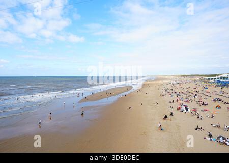Ein heller Sommertag an einem breiten Sandstrand, wo die Menschen schwimmen, sich sonnen und unter einem blauen Himmel mit weichen Wolken entlang der Küste spazieren. Stockfoto