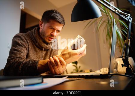 Mann, der am Schreibtisch sitzt und sein Handgelenk schmerzhaft hält, während er im Heimbüro an einem Laptop arbeitet. Das Konzept von Unwohlsein, Beanspruchung und Überbeanspruchung bei der Arbeit am Computer Stockfoto