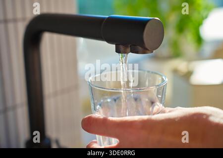 Hand halten Glas unter schwarzem Küchenarmatur mit Sonnenlicht vom Fenster in einem modernen Innenraum. Sauberes Wasser fließt vom Leitungsfilter in das Glas. Das Konzept des Trinkwassers und des Durstlöschens Stockfoto