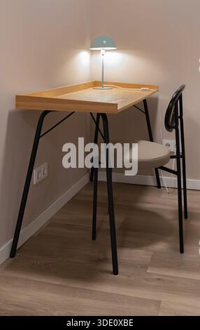 Horizontale Weitwinkelaufnahme eines eleganten Holzschreibtisches mit schwarzen Metallbeinen, einem Rattanstuhl und einer beleuchteten grünen Schreibtischleuchte in einer gemütlichen Zimmerecke Stockfoto