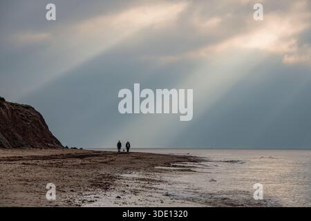 Zwei Personen spazieren entlang eines einsamen Winterstrandes an der Küste der isle of wight UK, wobei Sonnenstrahlen durch die Wolken brechen, die das Meer erleuchten. Stockfoto