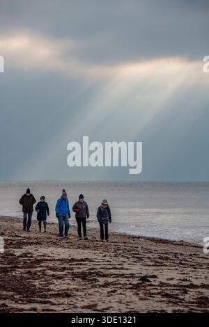 Gruppe von Menschen, die gemeinsam entlang eines einsamen Winterstrandes spazieren, mit Sonnenstrahlen und Lichtstrahlen, die durch die Wolken brechen und die Küste beleuchten. Stockfoto