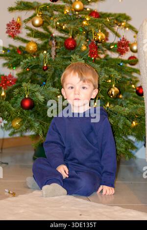 Kleinkind sitzt auf dem Boden vor einem geschmückten Weihnachtsbaum mit festlichen Lichtern in gemütlicher Atmosphäre. Stockfoto