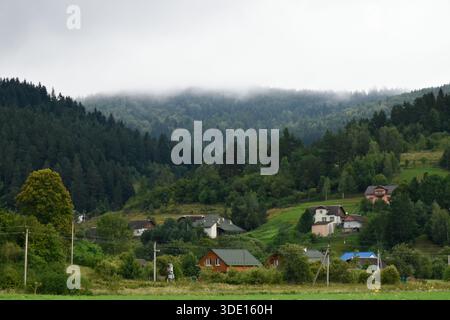Kleine farbenfrohe Häuser in Karpaten, ländliches Leben. Dicker Bergnebel in tiefen Kiefernwäldern, bewölkter Tag, stimmungsvolle Aussicht. Stockfoto