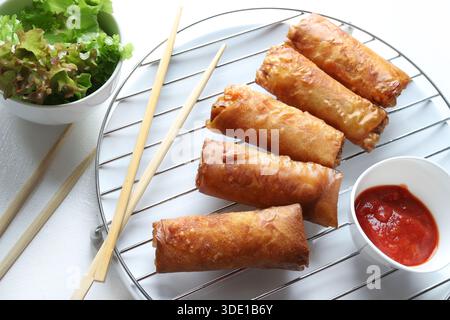 Knusprige gebratene Frühlingsrollen (chinesische Brötchen) serviert mit Essstäbchen, roter Sauce und frischem Salat auf weißem Hintergrund. Typisches Gericht der asiatischen Küche. Stockfoto