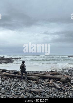 Ein Mann, von hinten gesehen, in einem Flanellhemd, stehend an einem Strand mit Treibholz und Felsen, im Winter an der Küste Oregons. Stockfoto