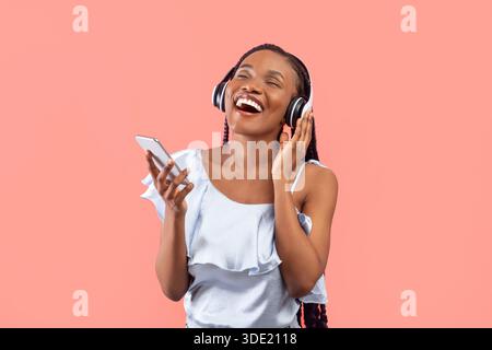 Junge schwarze Frau mit Kopfhörern und Smartphone, die Musik hört, die neue mobile App nutzt und die Augen vor Freude schließt Stockfoto