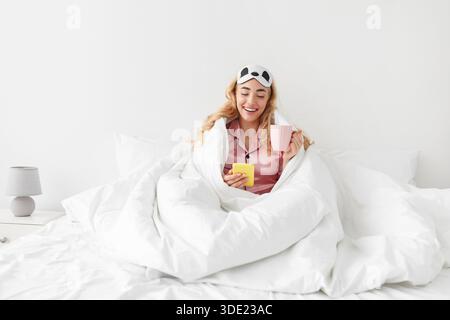 Lustige tausendjährige kaukasische blonde Frau in rosa Pyjamas und Schlafmaske genießt Kaffee und liest Nachricht auf dem Smartphone Stockfoto