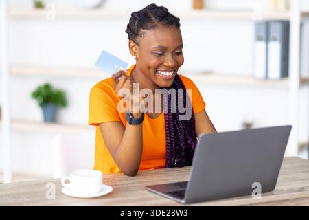Online-Vertrieb, E-Commerce. Fröhliche schwarze Frau, die mit Laptop und Kreditkarte am Tisch sitzt und zuhause im Internet einkaufen geht Stockfoto