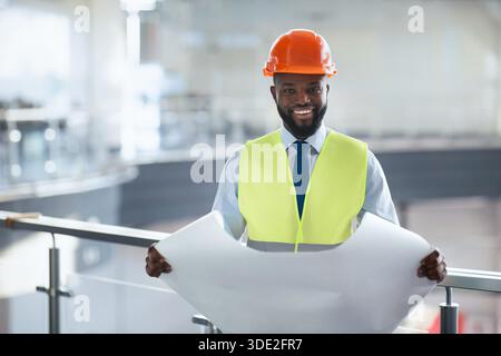 Bauarbeiter hält Baupläne auf einer modernen Baustelle mit Sicherheitsausrüstung und lächelndem Lächeln ab Stockfoto