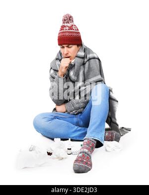 Kranker junger Mann im Winterhut hustet, während er mit Medikamenten und Taschentüchern auf dem Boden sitzt. Isoliert auf weißem Hintergrund. Hochwertige Fotos Stockfoto