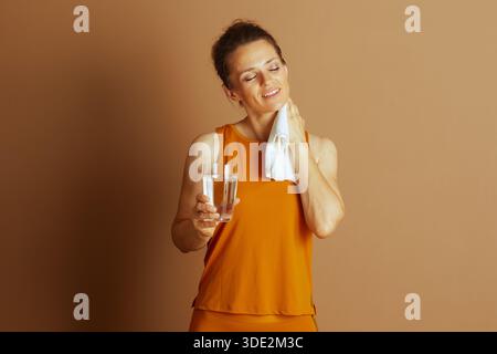 Eine Frau mittleren Alters in orangefarbener Aktivkleidung, mit geschlossenen Augen und einem ruhigen Lächeln hält Wasser und ein Handtuch. Sie verkörpert eine Erfrischung nach dem Workout Stockfoto