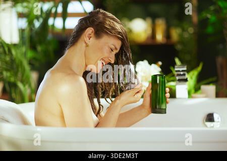 Lächelnde Frau mittleren Alters mit nassem, dunkelbraunem Haar in weißer Badewanne trägt das Produkt auf und hält eine grüne Flasche. Grüne Pflanzen und sanfte Beleuchtung sorgen für einen Stockfoto