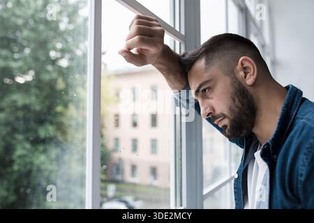 Trauriger, unglücklicher junger, gutaussehender Mann mit Hand auf Glas im Kücheninneren Stockfoto