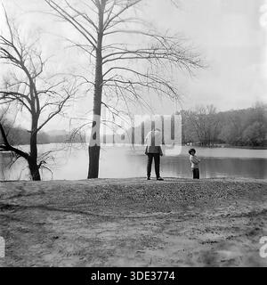 Ein monochromes Archivfoto aus der Sowjetzeit der 1980er Jahre zeigt Kravelis Dmitri Michailowitsch, der mit einer weiblichen Begleiterin am Uferufer in Brusino, Donezk, Ukraine UdSSR, steht. Der Mann, der eine Strickjacke trägt, blickt auf das ruhige Wasser der Seversky Donets, während die Frau neben ihm steht. Ein großer, blattloser Baum umrahmt die linke Seite der Komposition. Dieses malerische Bild dokumentiert einen ruhigen Moment der Freizeit im Freien und der Verbindung mit der Natur im friedlichen Donbass vor dem Krieg. Stockfoto