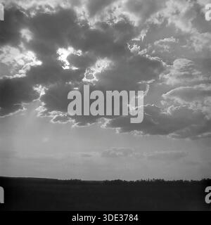 Das monochrome Archivfoto aus den 1970er Jahren der Sowjetzeit zeigt eine atemberaubende Skyscape über den offenen Steppen der Region Donezk, Ukraine UdSSR. Die Sonne verbirgt sich hinter massiven, dunklen Cumulus-Wolken, die einen dramatischen kontrastreichen Effekt mit hellen Silbereinfassungen und krepuskulären Strahlen erzeugen, die in Richtung des dunklen, niedrigen Horizonts strahlen. Dieses stimmungsvolle Bild unterstreicht die Weite des Himmels und die rohe, zeitlose Schönheit der ländlichen Landschaft im friedlichen Donbass vor dem Krieg und zeigt die Pracht der Natur. Stockfoto