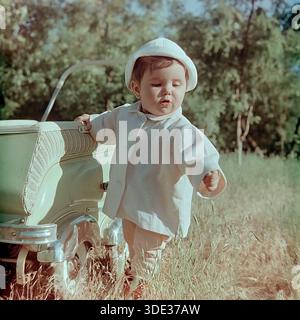Slawjansk, Region Donezk, Ukraine, UdSSR, Anfang der 1960er Jahre Ein nostalgisches Archivfoto eines niedlichen Kleinkindes, Inessa Mikhailovna Kravelis, in einem Sommerpark. Das Kind steht neben einem stilvollen hellgrünen Kinderwagen im Retro-Stil mit Chromdetails. Sie trägt einen weißen panamahut und einen leichten Anzug. Inessa schaut aufmerksam auf das Gras und erkundet die Welt um sie herum. Sonnenlicht erfüllt die Szene. Dieses authentische Bild fängt die Unschuld der Kindheit und die ruhige Atmosphäre des Familienlebens im friedlichen Donbass während der Sowjetzeit ein. Stockfoto