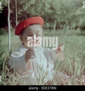 Slawjansk, Region Donezk, Ukraine, UdSSR, Anfang der 1960er Jahre Ein wunderbares Archivfoto in Vintage-Farben von Inessa Mikhailovna Kravelis, einem niedlichen Kleinkind, das auf hohem Gras in einem sonnigen Park sitzt. Das Kind trägt ein hellweißes Sommeroutfit und eine auffällige rote Baskenmütze. Sie schnickt die Augen fest, macht ein lustiges und entzückendes Gesicht, vielleicht reagiert sie auf die helle Sonne. Im Hintergrund sind grüne Bäume zu sehen. Dieses ehrliche Bild fängt die Unschuld, Freude und sorglose Atmosphäre der Kindheit im friedlichen Donbass während der Sowjetzeit ein. Stockfoto