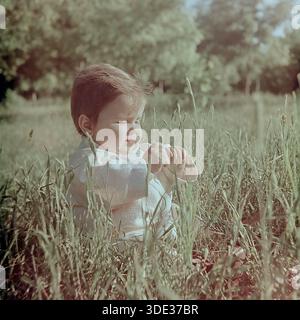 Slawjansk, Region Donezk, Ukraine, UdSSR, Anfang der 1960er Jahre Ein wunderschönes Archivfoto in Vintage-Farben von einem niedlichen Kleinkind, Inessa Mikhailovna Kravelis, das in hohem Gras in einem sonnigen Park sitzt. Das Kind trägt ein hellweißes Sommeroutfit ohne Hut. Sie hält ihre Hände zusammen, blickt mit einem konzentrierten und ernsten Ausdruck nach unten und spielt vielleicht mit einem Grashalm. Sonnenlicht beleuchtet ihr kurzes Haar. Dieses authentische Bild fängt die Unschuld, Gelassenheit und glückliche Atmosphäre der Kindheit im friedlichen Donbass während der Sowjetzeit ein. Stockfoto
