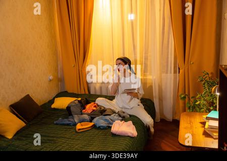Junge kaukasische Frau organisiert Kleidung in einem leise beleuchteten Raum bei Sonnenuntergang. Sie faltet Kleidung ordentlich. Design für sauberes Zuhause und organisierte Räume. Weitsicht. Stockfoto