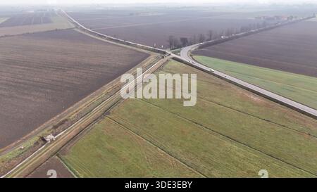 Ein ausgedehnter Drohnenschuss erfasst die geometrischen Muster von bewirtschaftetem Land, das an einer Kreuzung auf eine gewundene Asphaltstraße trifft, wobei Fahrzeuge navigieren Stockfoto