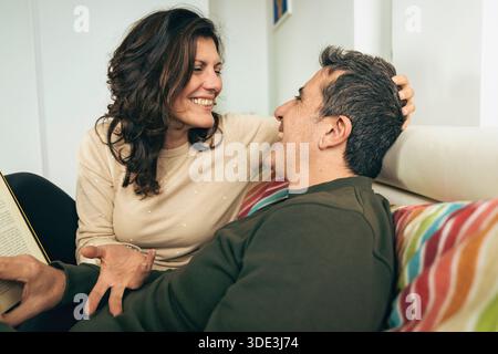 Authentisches Paar mittleren Alters, das einen lustigen Moment beim Lesen im Bett teilt. Die Frau berührt liebevoll die Haare des Ehemanns. Das Konzept der glücklichen Ehe und c Stockfoto