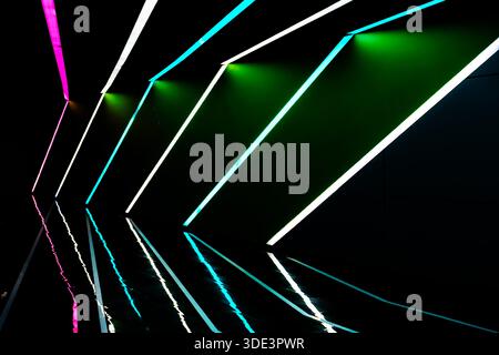 Eine moderne, abstrakte Lichtinstallation, neonfarbene Lichtstreifen sind diagonal vor dunklem Hintergrund angeordnet. Die Lichter reflektieren lebhaft auf einem gl Stockfoto